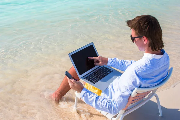 Młody biznesmen za pomocą laptopa i telefon na tropikalnej plaży — Zdjęcie stockowe