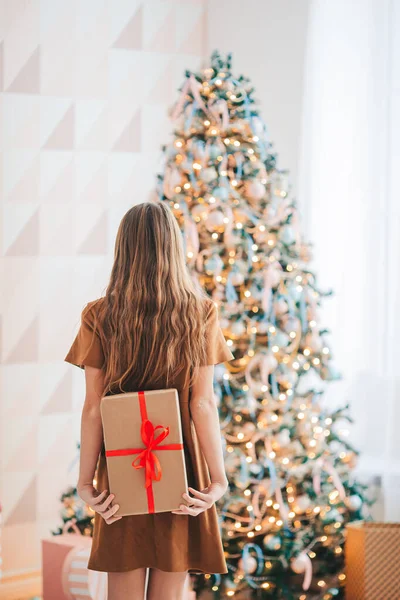 Visão traseira da criança segura uma caixa de presente perto da árvore de Natal dentro de casa. — Fotografia de Stock