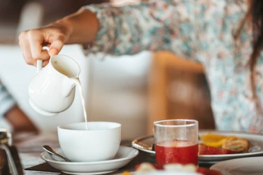 Woman pouring milk in mug from kettle. Breakfast time