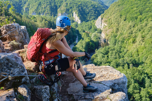 Woman climber with helmet and backpack sits down and relaxes at the end of a via ferrata in Suncuius, Romania, gazing towards Crisul Repede defile covered with green forests, in the morning light.