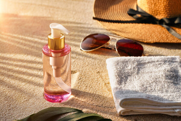 Sunglasses near straw hat and bottle with suntan oil on sandy beach, towel and shadow from palm leaves