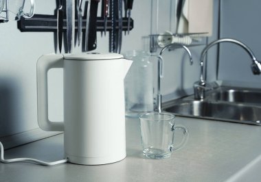 Elektrikli su ısıtıcısı beyaz mat yüzeyi, mutfak parçası. Arka planda filtrelenmiş su bardağı ve musluğu