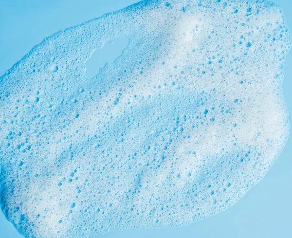 Face cleansing mousse sample. White cleanser foam bubbles on blue background. Soap, shower gel, shampoo foam texture closeup.