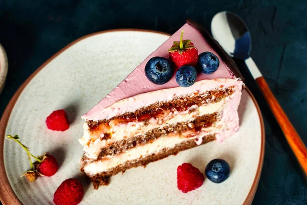 A piece of cake, chocolate biscuit with cream cheese filling on dark blue background close up. Berries on pink cream decoration. — Stock Photo, Image