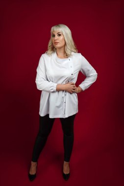 Kendine güvenen, genç, sarı saçlı kadın kırmızı arka planda dikilirken kameraya bakıyor.