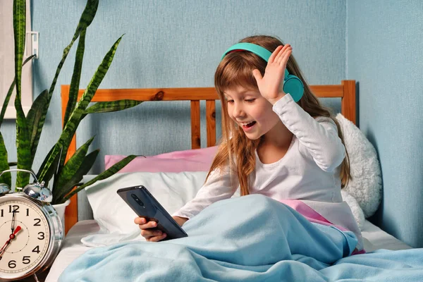 stock image Happy cute little vlogger waving hand saying hello hi looking at camera talking to webcam, smiling kid child girl making online video call recording vlog sitting on bed at home, portrait