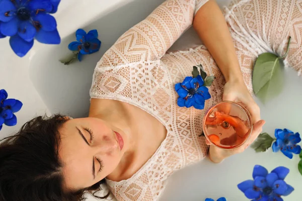 Güzellik mankeni kız bir bardak gül şarabı, spa ve cilt bakımı konseptiyle süt banyosu yapıyor. Mavi çiçekli güzel bayan süt banyosunda dinleniyor. Gençleştirme. — Stok fotoğraf