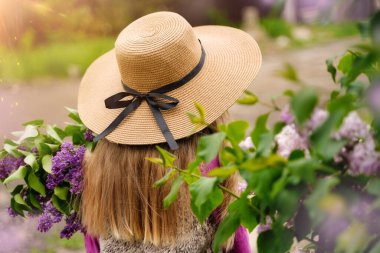 Hasır şapkalı, uzun sarı saçlı, bahar günü takılan küçük bir kız. Çiçekli leylak çalıları. Yüksek kalite fotoğraf