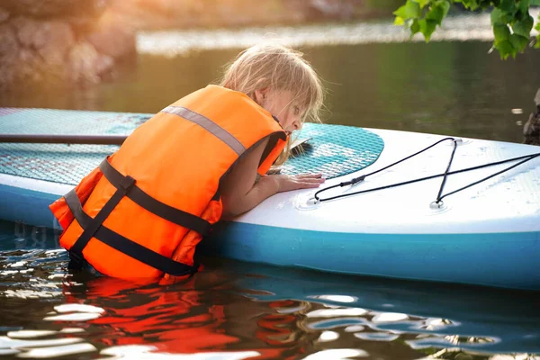 European child girl in a life jacket climbs on a SUP board floating on the lake. High quality photo