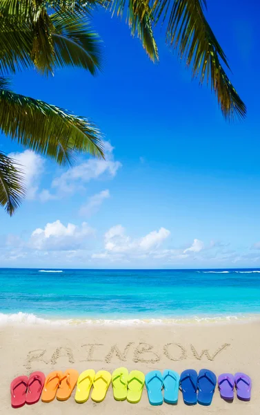 Надпись "Радуга" и цветные шлепанцы на песчаном пляже — стоковое фото