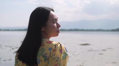 Güneşli bir günde göl kenarında manzaranın keyfini çıkaran Asyalı kadın turist. Açık hava seyahati ve doğa teması.