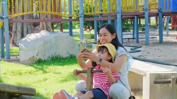 年轻妈妈拿着智能手机给女儿在户外操场上玩电锯的照片 快乐的小女孩 妈妈在孩子们的操场上笑着 — 图库视频影像