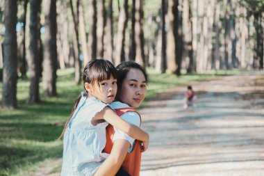 Parkta annesiyle sırt sırta gezintiye çıkmış sevimli genç bir kız. Anne kızı sırtında tutuyor ve yaz bahçesinde yürüyüşe çıkıyor..