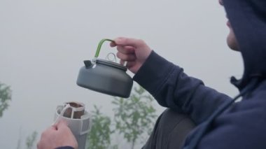 Genç turistlerin yakın çekim elleri dağın zirvesinde kahve ile sıcak suyu bardağa döküyor. Genç bir turist bir bardaktan sıcak bir içecek içer ve dağlardaki manzaranın tadını çıkarır..