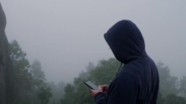 Lacivert erkek kapüşonlu uzun kollu kazak giyen genç adam akıllı telefon kullanıyor ve arka planda kalın bir sis olan dağların manzarasının tadını çıkarıyor..