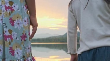 Anne, yaz günü gün batımında göl kenarında kızıyla el ele tutuşurken. Mutlu aile tatillerinde birlikte vakit geçiriyor..