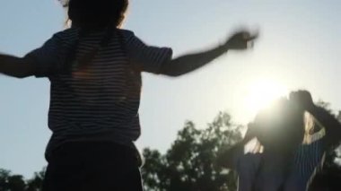 Silhouette of happy sisters raising their hands and jumping in the summer garden at sunset. Cheerful girls jumping and playing in the park.