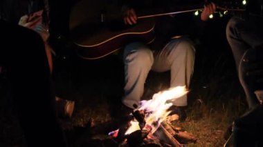 Neşeli genç arkadaşlar parlak bir kamp ateşinin etrafında oturur, gitar çalar ve gölün kenarında şarkı söylerler..