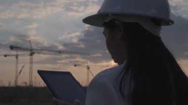 Gün batımında inşaat alanında tablet bilgisayarlı kadın inşaat mühendisi. Beyaz kasklı, kendine güvenen bir mimar inşaat alanına bakıyor..