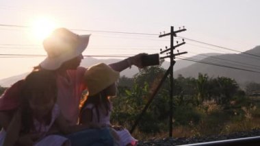 Asyalı anne ve çocuk tren raylarında oturur ve gün batımında akıllı telefonlarla selfie çekerler..