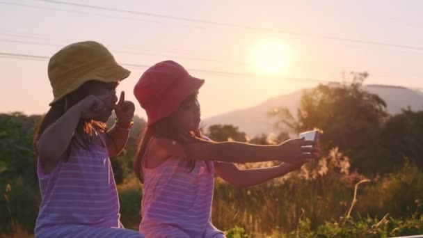 2人の姉妹はトラックに座って 日没時にスマートフォンと一緒に自撮りします アジアの姉妹は休暇で一緒に過ごすのを楽しむ — ストック動画