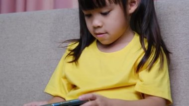Evdeki kanepede oturmuş akıllı telefonuyla oyun oynayan sevimli küçük kız. Modern iletişim kavramları ve alet bağımlılığı