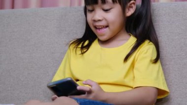 Evdeki kanepede oturmuş akıllı telefonuyla oyun oynayan sevimli küçük kız. Modern iletişim kavramları ve alet bağımlılığı