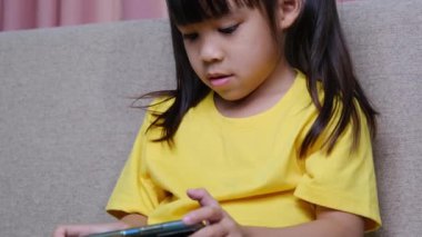 Evdeki kanepede oturmuş akıllı telefonuyla oyun oynayan sevimli küçük kız. Modern iletişim kavramları ve alet bağımlılığı