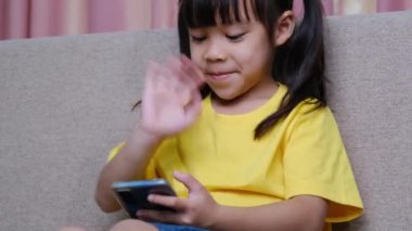 Sevimli küçük kız video araması yapıyor ve akıllı telefonuyla selamlarını sallıyor. Evdeki kanepede oturuyor..