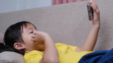 Sevimli küçük kız video araması yapıyor ve akıllı telefonuyla selamlarını sallıyor. Evdeki kanepede yatıyor..