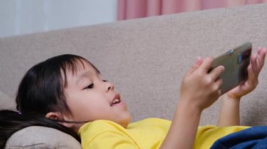 Evdeki kanepede uzanmış akıllı telefonuyla online oyunlar oynayan sevimli küçük kız. Modern iletişim kavramları ve alet bağımlılığı
