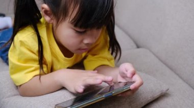 Evdeki kanepede uzanmış akıllı telefonuyla online oyunlar oynayan sevimli küçük kız. Modern iletişim kavramları ve alet bağımlılığı