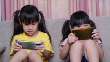 İki kız kardeş evdeki kanepede oturmuş akıllı telefonlarıyla oyun oynuyorlar. Modern iletişim ve alet bağımlılığı kavramı. Elinde aletler olan iki çocuk..