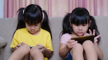 İki kız kardeş evdeki kanepede oturmuş akıllı telefonlarıyla oyun oynuyorlar. Modern iletişim ve alet bağımlılığı kavramı. Elinde aletler olan iki çocuk..