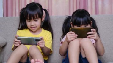 İki kız kardeş evdeki kanepede oturmuş akıllı telefonlarıyla oyun oynuyorlar. Modern iletişim ve alet bağımlılığı kavramı. Elinde aletler olan iki çocuk..