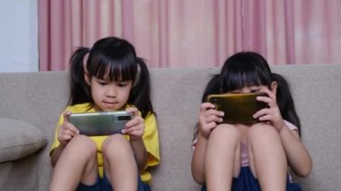İki kız kardeş evdeki kanepede oturmuş akıllı telefonlarıyla oyun oynuyorlar. Modern iletişim ve alet bağımlılığı kavramı. Elinde aletler olan iki çocuk..