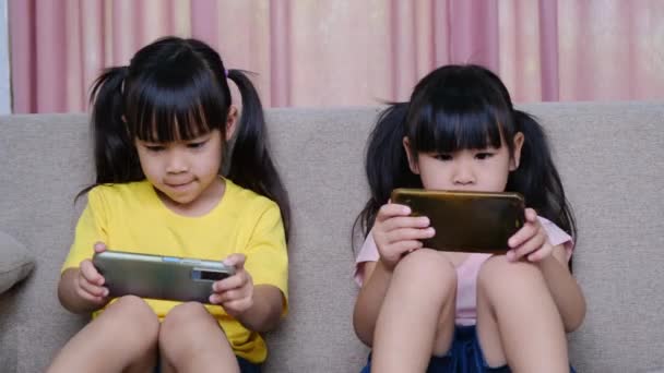 两个姐姐坐在家里的沙发上 用她们的智能手机玩网络游戏 现代通信和小玩意成瘾的概念 两个带小玩意的孩子 — 图库视频影像