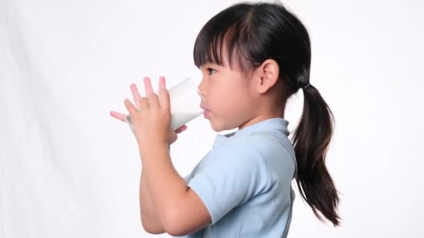 アジア系のかわいい女の子は ガラスから牛乳を飲んで スタジオで白い背景に彼女の唇をなめる 小さな子供のための健康的な栄養 — ストック動画