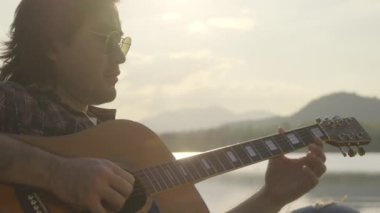 Yakışıklı kıvırcık saçlı Asyalı adam gün batımında göl kenarında gitar çalıp şarkı söylüyor..