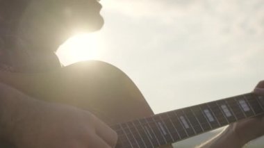 Yakışıklı kıvırcık saçlı Asyalı adam gün batımında göl kenarında gitar çalıp şarkı söylüyor..