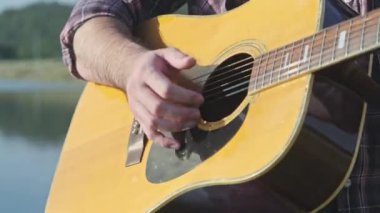 Yakışıklı kıvırcık saçlı Asyalı adam gün batımında göl kenarında gitar çalıp şarkı söylüyor..