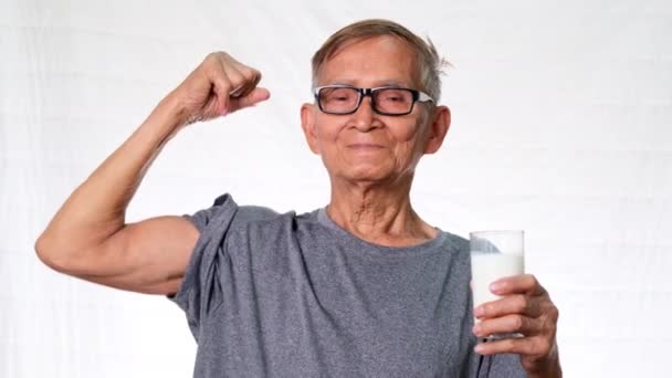 健康的老人一边举着一杯牛奶 一边展示自己的肌肉 自豪地微笑在工作室的白色背景上 — 图库视频影像