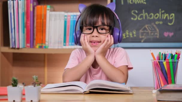可爱的小女孩 带着耳机听着桌上放着英语学习书的有声读物 学习英语和现代教育 — 图库视频影像
