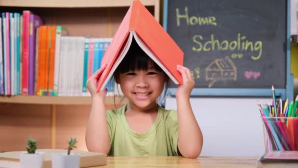 かわいい女の子の屋根のように彼女の頭の上に本を保持し 笑顔とカメラを見て 家庭教育のための愛らしい子供読書本 — ストック動画