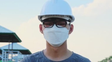 Miğfer ve maske takan genç Asyalı mühendis arka plandaki kameraya bakıp gülümsüyor. Coronavirus salgını sırasında bir barajda çalışan genç bir mühendis..