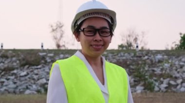 Kendine güvenen kadın mühendis kollarını kavuşturup kameraya gülümsüyor. Rezervuarın taş duvar arka planına karşı..