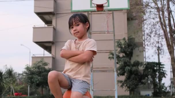 かわいい女の子はバスケットボールに座って 熱心に見て 屋外のバスケットボールコートで笑顔 — ストック動画
