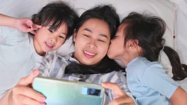 快乐的亚洲家庭享受与智能手机舒适的家 用电话微笑着妈妈和可爱的女儿 在床上用自拍或视频剪辑来回忆 在家里玩的开心 — 图库视频影像