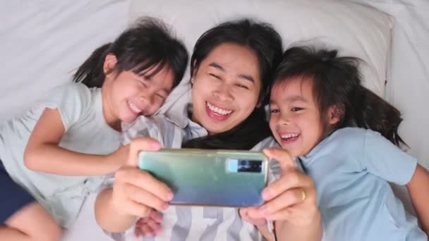 快乐的亚洲家庭享受与智能手机舒适的家 用电话微笑着妈妈和可爱的女儿 在床上用自拍或视频剪辑来回忆 在家里玩的开心 — 图库视频影像
