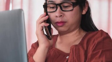 Günlük kıyafetler içinde güzel bir Asyalı kadın telefonla konuşuyor ve bilgisayarını kullanıyor. Ofis masasında oturuyor. Ofiste yalnız çalışan meşgul bir iş kadını..